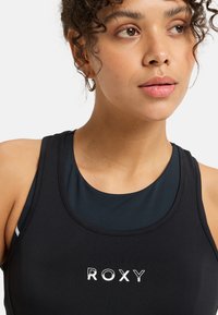 Black sports tank top with a layered design and a round neckline. Features a white logo "ROXY" on the front and smooth, fitted fabric.