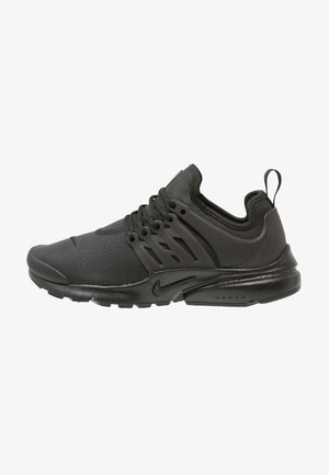 Zapatillas deportivas negras con un upper texturizado, suela curva y cordones trenzados. El lateral presenta múltiples cortes de ventilación y una pestaña en el talón.