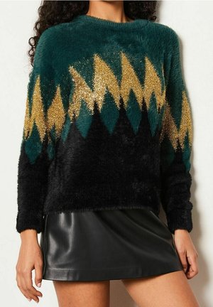 Vrouw die een zachte trui draagt met een groene en zwarte basis en een gouden zigzagpatroon, gecombineerd met een korte zwarte leren rok.