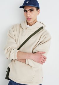 Beige fleece hettegenser med snor, har en frontlomme, brukt med en grønn crossover stropp. Mørkeblå denimjeans og blå caps fullfører looken.