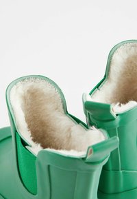Botas de goma verdes con forro de felpa beige, vistas desde un ángulo superior sobre un fondo blanco.