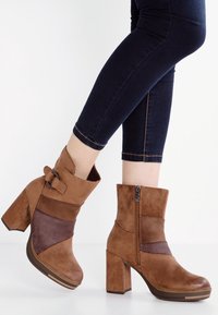 Bottines en suède marron avec un talon bloc, présentant une fermeture éclair et une sangle décorative. Design à panneaux avec des nuances de brun variées.