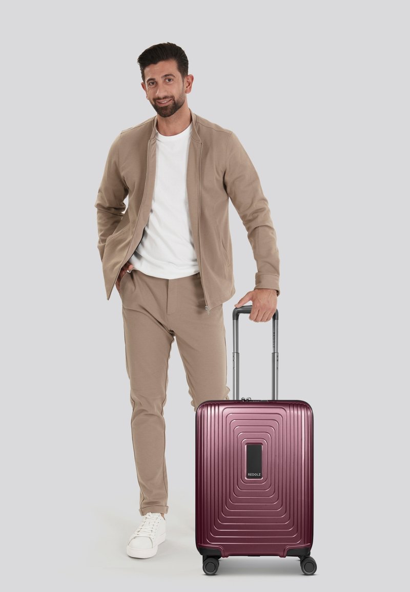 Valigia rigida di colore bordeaux lucido con texture lineare, maniglia integrata e quattro ruote. Uomo in abbigliamento beige si trova accanto ad essa.