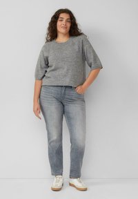 Kobieta z kręconymi włosami, ubrana w szary sweter z krótkim rękawem, wyblakłe szare dżinsy i białe trampki, stoi z jedną ręką w kieszeni.