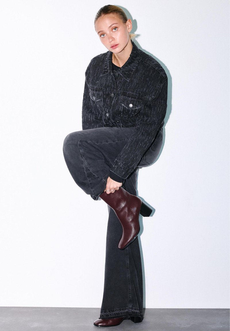 Woman in dark denim jacket and jeans adjusts one burgundy heeled ankle boot against a white background.