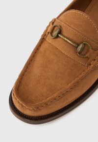 Καφέ μοκασίνι από σουέντ με διακοσμητικό χυτό μπρούτζινο στοιχείο σε σχήμα αλόγου και εμφανείς ραφές σε σκούρα σόλα, απεικονισμένο σε λευκό φόντο.