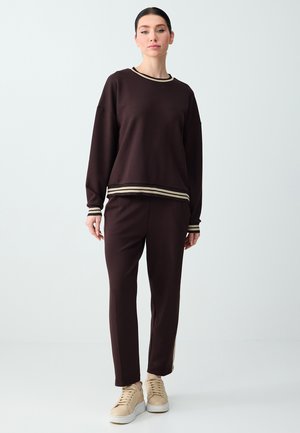 Femme portant un haut à manches longues marron foncé et un pantalon assorti avec des poignets à rayures beiges et des baskets beiges, debout devant un fond uni.