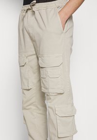 Béžové kalhoty typu cargo vyrobené z bavlny s elastickým pasem, stahovací šňůrkou a několika velkými kapsami po stranách pro uložení věcí.