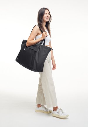 Glimlachende vrouw die een wit cropped topje en wijde crème broek draagt, met een grote zwarte totebag over haar schouder, lopend naar rechts.