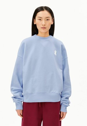 Persoon met lang donker haar draagt een oversized lichtblauwe trui en donkerrode broek, staande tegen een effen witte achtergrond.