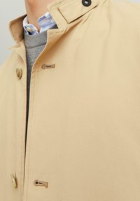 Beige buitenjack met een hoge kraag, voorzien van een knoopsluiting, textuurstof en een kleine borduursel detail nabij de kraag.
