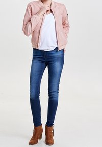 Person som bär en ljusrosa bomberjacka, vit t-shirt, blå skinny jeans och bruna mocka ankelskor, står med ena handen i fickan.