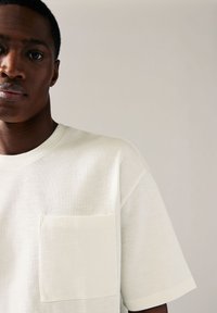 Weißes oversized T-Shirt mit Brusttasche, Rundhalsausschnitt und kurzen Ärmeln. Der Stoff hat eine strukturierte Oberfläche. Schlichtes Design.