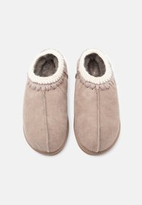 Ett par beige mockaskor med vit stickad kant och mjukt fleecefoder, sett uppifrån mot en neutral bakgrund.