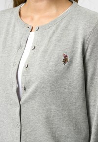 Grijze gebreide cardigan met een ronde hals, knoopsluiting en een klein geborduurd ontwerp op de linkerkant van de borst. Soepele textuur met gemiddeld gewicht.
