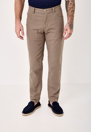 Mann trägt beige, gerade geschnittene Hose, marineblaue Slip-On-Schuhe, marineblaues Hemd, steht mit tätowierten Armen entspannt an den Seiten.