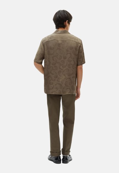 Camicia marrone a maniche corte con un sottile motivo paisley, progettata con una vestibilità rilassata, abbinata a pantaloni olive a gamba affusolata e scarpe nere.