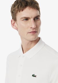 Hvid polo skjorte med knap-til-krave, med et lille grønt krokodillelogo på brystet, lavet af glat stof og let struktureret.