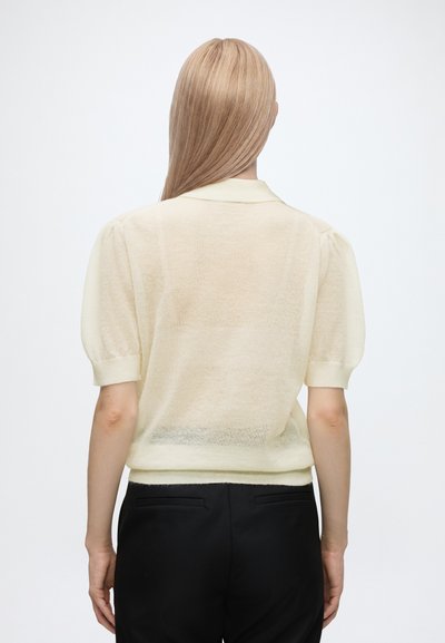Personne blonde aux cheveux raides portant un pull crème à manches courtes et un pantalon noir, vue de dos sur fond blanc.
