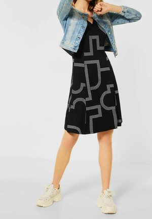 Frau trägt ein schwarzes Kleid mit weißen geometrischen Mustern, eine hellblaue Jeansjacke und weiße klobige Sneaker, steht mit erhobenen Armen.