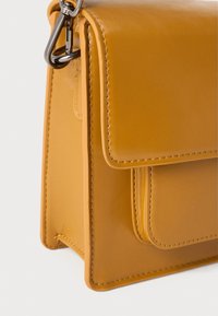 Sac à main en cuir jaune avec une texture lisse. Comprend une fermeture à rabat, une poche latérale et des attaches en métal argenté. Forme rectangulaire compacte.