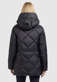 Femme portant une veste rembourrée matelassée noire avec capuche, vue de dos, se tenant devant un fond gris uni.