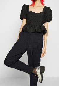 Svart texturerad peplumtop med korta puffärmar, kombinerad med mörka rutiga byxor. Skorna består av glänsande silvriga loafers och mönstrade strumpor.