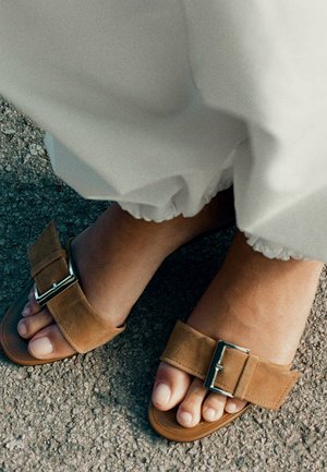 Pieds dans des sandales en daim beige avec boucles métalliques portées avec un pantalon blanc froncé, debout sur un sol en asphalte.