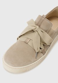 Beige semsket sneaker med en teksturert stoffsløyfe, en polstret såle og en rillede gummisåle for grep. Minimalistisk design med synlige detaljer.