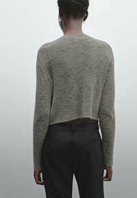 Henkilö, jolla on päällään harmaa lyhytneule ja mustat housut, nähtynä takaa vaaleanharmaata taustaa vasten.