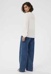Femme avec des cheveux bouclés courts portant une blouse blanche transparente à manches longues et un jean bleu à jambes larges, se tenant debout et regardant par-dessus son épaule.