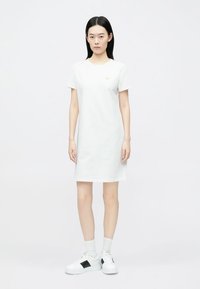White cotton t-shirt dress with short sleeves, a round neckline, and a straight cut. Worn with white sneakers featuring black accents.