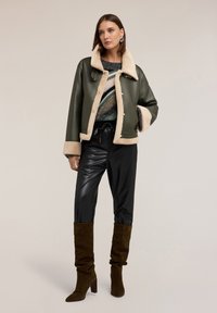 Kobieta w oliwkowej, jagnięcej kurtce, wielokolorowym swetrze dzianinowym, czarnych skórzanych spodniach i brązowych zamszowych kozakach do kolan, stojąca z ręką w kieszeni.