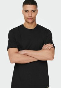 Schwarzes Kurzarm-T-Shirt aus glattem Baumwollstoff, mit rundem Ausschnitt und normalem Schnitt, getragen von einer Person mit verschränkten Armen.