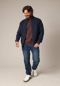 Marine blauwe zip-up jas met staande kraag, gecombineerd met een bruine t-shirt, blauwe spijkerbroek en witte sneakers. Voorzien van zijzakken en geribbelde manchetten.
