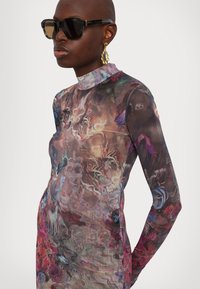 Robe à manches longues avec des motifs abstraits vibrants dans des tons de marron, rose et bleu ; col montant et coupe ajustée. Lunettes de soleil surdimensionnées noires et boucles d'oreilles dorées.