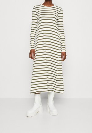 Žena v midi šatech s dlouhými rukávy v bílé a olivově zelené pruhované barvě, obutá do bílých robustních kotníkových bot na platformě, stojící před jednobarevným pozadím.