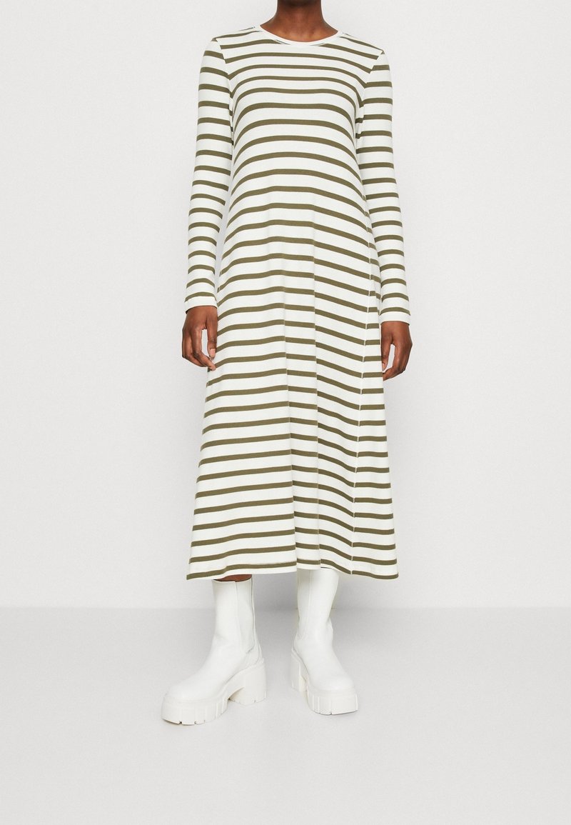 Femme portant une robe midi à manches longues rayée blanche et vert olive, avec des bottines blanches à plateforme épaisse, debout devant un fond uni.