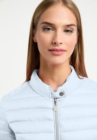 Jasnoniebieska kurtka puchowa o pionowej tłoczonej teksturze, wysoki kołnierz oraz zamek błyskawiczny z okrągłym metalowym zawieszką.