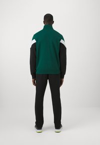 Tuta verde e nera, caratterizzata da un alto colletto, accentuazioni bianche a contrasto sulle spalle e un design aderente. Tessuto morbido, look sportivo.