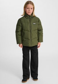 Barn med axellångt blont hår som bär en olivgrön pufferjacka, svarta byxor och svarta sneakers med bruna sulor, stående.