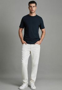 Tricou bleumarin închis cu guler rotund, pantaloni din bumbac albi și adidași albi. Modelul stă cu mâinile în buzunare pe un fond neutru.