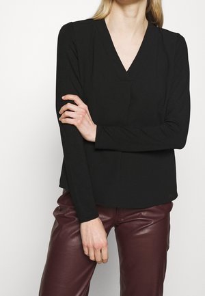 Frau trägt eine schwarze, langärmelige V-Ausschnitt-Bluse und dunkelrote Lederhose, mit einem Arm verschränkt und dem anderen locker herabhängend.