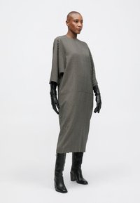 Egy kopasz nőt látunk, aki hosszú, laza szürke ruhát, fekete bőrvédő kesztyűt és térd fölé érő fekete bőrcipőt visel, fehér háttér előtt áll.