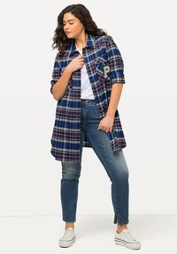 Blauw en rood geruite overhemdenjack met knoopsluiting, zijzakken en een letterschild, gecombineerd met lichtblauwe jeans en witte sneakers.