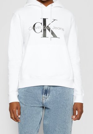 White hoodie with front pocket and Calvin Klein Jeans logo in black and gray, worn with light blue denim jeans.
