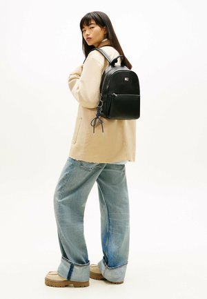 Giovane donna con giacca beige e jeans larghi che indossa uno zaino in pelle nero con portachiavi, in piedi davanti a uno sfondo bianco uniforme.