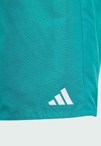 Tyrkysové atletické šortky z ľahkého materiálu, s bielym logom Adidas v trojuholníkovom dizajne na dolnom ľavom rohu. Hladká textúra.