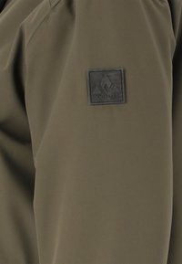 Ärmel einer olivgrünen Jacke mit einem schwarzen quadratischen Aufnäher, der ein Berglogo und das Wort "WHISTLER" zeigt.