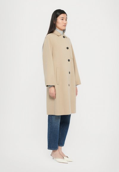 Beige wool coat with a button front, large collar, and side pockets, paired with blue jeans and white slip-on shoes, standing on a plain background.